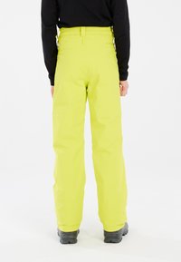 Gele waterdichte skibroek met een rechte snit, voorzien van een hoge taille en verstelbare banden aan de achterkant. Zwarte schoenen zichtbaar eronder.