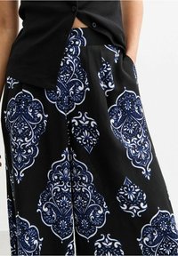 Černé kalhoty s širokými nohavicemi, které mají vzor v bílé a námořnické modři, hladkou texturu a kapsy po stranách.