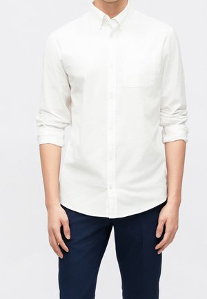 Homme portant une chemise blanche boutonnée avec les manches retroussées et un pantalon bleu marine devant un fond clair uni.