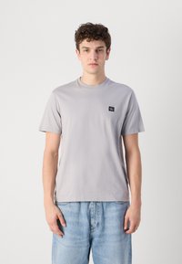 Camiseta gris clara de algodón suave, con cuello redondo y mangas cortas. Pequeño emblema en el lado izquierdo del pecho. Combinada con jeans de denim.