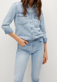 Mujer vistiendo una camisa de denim azul claro con mangas arremangadas y jeans de denim azul claro a juego, con una mano en el bolsillo.