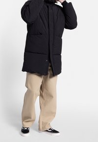 Schwarzer Steppmantel mit hohem Kragen, Druckknöpfen und gesteppter Optik, kombiniert mit beigen Hosen mit weitem Bein und schwarzen Sneakers.