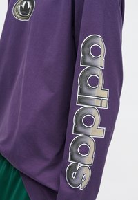 Violeta garām piedurknēm krekls ar apaļu logotipu ar lapu dizainu un "adidas" tekstu biezā, gradientā tipogrāfijā gar piedurkni.