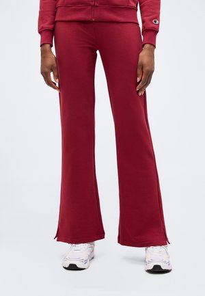 Person iført vinrøde vide sweatpants, matchende hættetrøje med lynlås og hvide sneakers, stående mod en ensfarvet baggrund.