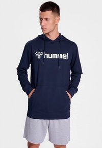 Granatowa bluza z kapturem z przednią kieszenią i sznurkami, z dużym białym logo. Do tego jasnoszare szorty. Gładka struktura tkaniny.