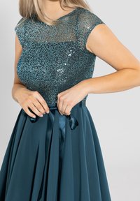 Robe de soirée en sarcelle avec un bodice pailleté et une jupe fluide. Un ruban satiné noué à la taille ajoute une touche de détail. Manches courtes, tissu texturé.