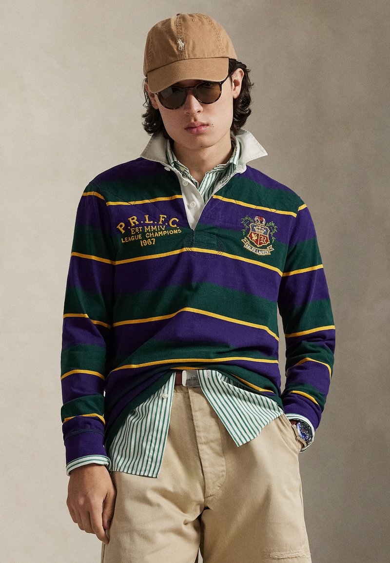 Maillot de rugby rayé violet et vert avec des accents jaunes, col blanc et blason brodé, porté sur une chemise rayée verte, pantalon beige.