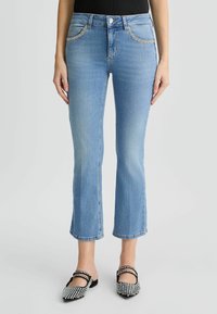 Jean en denim bleu clair à coupe évasée, avec poches avant et couture décorative. Portés avec des ballerines noires et blanches ornées.