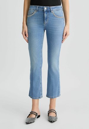 Jean en denim bleu clair à coupe évasée, avec poches avant et couture décorative. Portés avec des ballerines noires et blanches ornées.