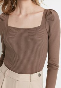 Braunes, geripptes Langarmshirt mit quadratischem Ausschnitt und gepufften Schultern, kombiniert mit beigen, hoch taillierten Hosen mit Knopfdetail.