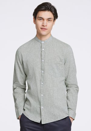 Camisa larga de manga larga verde, texturizada, con cuello mandarín, cierre frontal de botones y un bolsillo en el pecho. La tela parece ligera.
