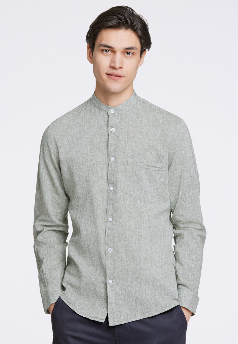 Camicia verde a maniche lunghe con texture, colletto alla mandarino, chiusura con bottoni frontali e taschino sul petto. Il tessuto sembra leggero.