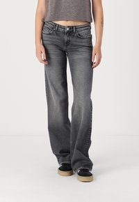 Personne portant un jean noir délavé coupe bootcut, des chaussures noires avec semelles beige, et un top gris court, debout devant un fond blanc uni.