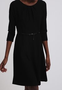 Vestido negro texturizado con mangas de tres cuartos, escote redondo plisado y un cinturón negro delgado en la cintura.