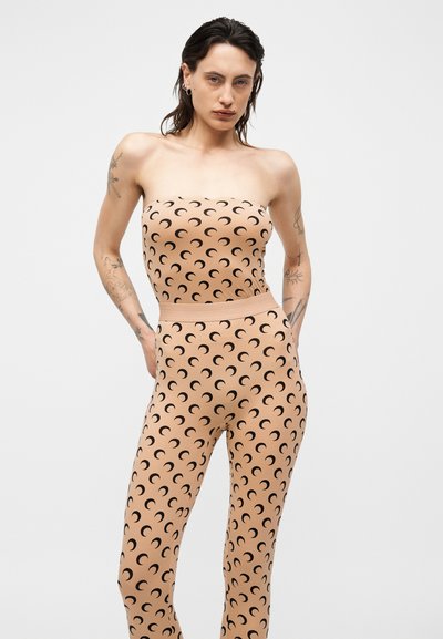 Ženska v bež brezramnem kombinezonu z motivom črnih rdečih polmesecov, stoji na enobarvni svetlo sivi podlagi.