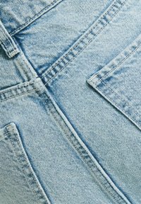 Ljusblå denimtyg med en framträdande textur, med synliga sömmar och snedställda fickdesigner. Kentvävsmönster är tydligt.