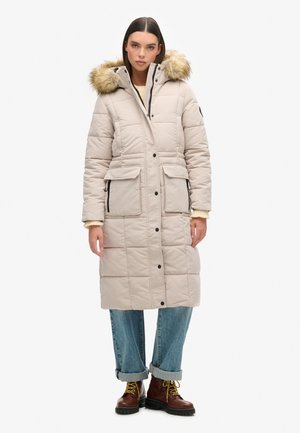 Long manteau matelassé beige avec capuche en fourrure, fermeture éclair à l'avant, deux poches sur la poitrine et deux poches sur les côtés, porté par-dessus un jean retroussé et des bottes.