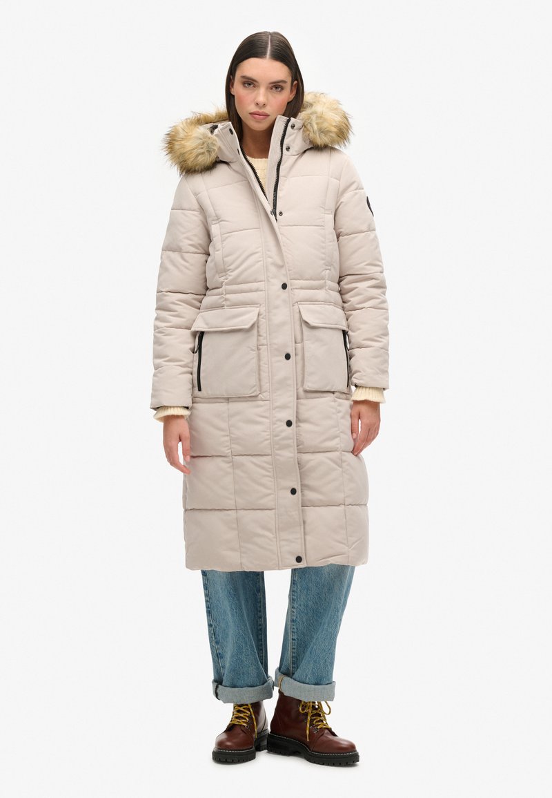 Lang beige pufferjakke med pelsforet hætte, lynlås foran, to brystlommer og to sidelommer, båret over oprullede jeans og støvler.