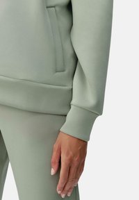 Felpa verde chiaro con un design elegante, texture morbida e una tasca laterale, con un pantalone abbinato visibile.