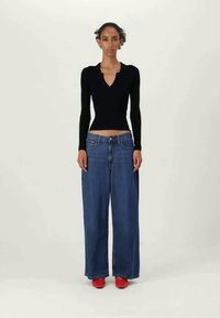 Nő áll, testre simuló fekete hosszú ujjú felsőt, bő szárú kék farmert és piros cipőt visel egy sima világos háttér előtt.