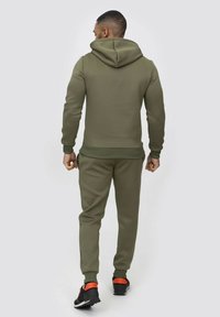 Ensemble en vert olive composé d'un hoodie et d'un pantalon de survêtement, doté d'une capuche à cordons, de poignets côtelés et d'une texture lisse. Chaussures de sport noires et oranges.