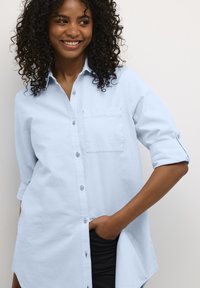 Vaaleansininen napitettava paita, jossa on tasku, käännetyt hihat ja hieman kaartuva helma. Pehmeä koostumus, rento muotoilu.