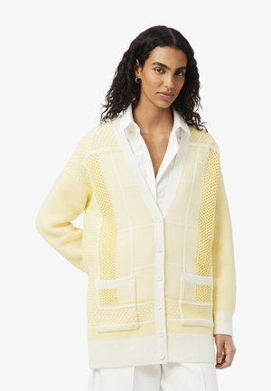 Mujer con cabello oscuro y rizado que lleva una camisa blanca y un cárdigan tejido de color amarillo pálido con grandes bolsillos delanteros, de pie frente a un fondo blanco.
