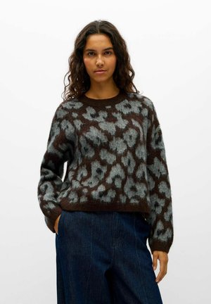 Une femme porte un pull à motifs marron et gris avec un jean bleu foncé, sur un fond blanc uni.