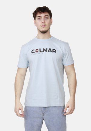 Giovane uomo che indossa una t-shirt Colmar Originals azzurro chiaro e jeans azzurro chiaro, in piedi con le braccia leggermente piegate contro uno sfondo neutro.