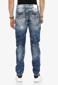 Blaue Jeans aus Denim mit einem verwaschenen Finish, die ein einzigartiges gestepptes Design auf den Gesäßtaschen und eine schmal zulaufende Passform aufweisen. Kombiniert mit weißen Turnschuhen.