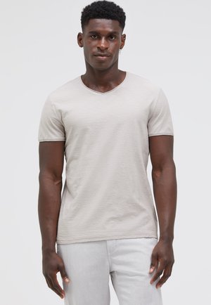 Man with short curly hair wearing a light beige V-neck T-shirt and light-colored pants, standing against a plain background.