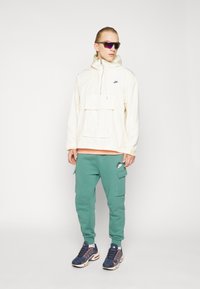 Lys beige pullover jakke med forlomme, parret med grøn cargo bukser. Tilbehøret inkluderer mørke solbriller og marineblå sko til sport med orange detaljer.