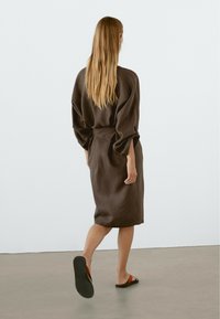 Robe en lin marron avec une coupe décontractée, longueur mi-mollet, manches larges et ceinture nouée. Associée à des sandales plates.