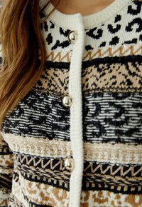 Strickjacke mit einer Mischung aus beigen, schwarzen und cremefarbenen Mustern. Silberne Knöpfe, strukturierte Wolle und ein runder Ausschnitt mit geometrischen Akzenten.