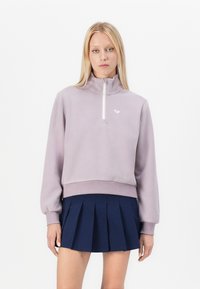 Woman wearing a lavender half-zip fleece pullover and a navy blue pleated skirt against a plain white background.