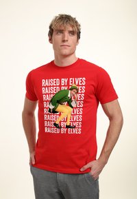 Červené bavlněné tričko s opakovaným textem "RAISED BY ELVES" a barevným grafickým zobrazením elfa ve světlých zelených šatech se žlutými nohama.