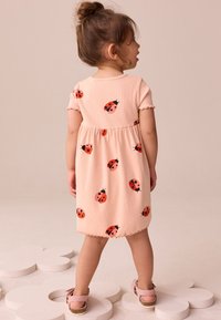 Palen rosa klänning med korta ärmar och en markerad midja, prydd med röda nyckelpigemönster. Klänningen har en vågig fåll och texturerat tyg.