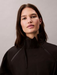 Black waterproof pullover jacket with high collar, featuring zippers and a subtle embossed logo on the front. Smooth, matte texture.