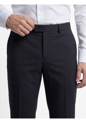 Mann befestigt einen Knopf an schwarzen formellen Hosen, trägt ein weißes Hemd mit langen Ärmeln, vor einem einfarbigen weißen Hintergrund.