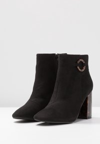 Botines de ante negros con punta afilada y un tacón grueso con patrón de carey. Presentan una cremallera lateral y herrajes circulares decorativos.