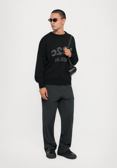 Sort striktrøje med logo, mørke bukser og sorte sneakers. Accessories inkluderer mørke solbriller og en lille taske med rem.