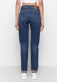 Femme portant un jean droit bleu foncé retroussé, un top court blanc et des baskets blanches, debout dos à la caméra sur un fond uni.