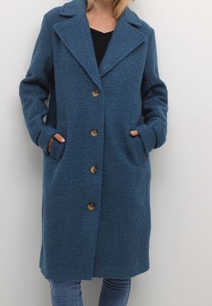 Femme portant un long manteau bleu texturé avec des revers et quatre boutons à l'avant, les mains dans les poches, associé à un jean bleu et un haut noir.