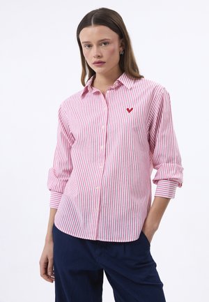 Jeune femme portant une chemise à rayures roses et blanches avec un petit cœur rouge sur la poitrine, associée à un pantalon bleu foncé.