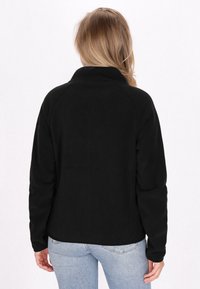 Musta fleece-takki korkealla kauluksella ja raglan-hihoilla. Pehmeä rakenne, minimaalinen yksityiskohtaisuus ja istuva siluetti, yhdistettynä vaaleansinisiin farkkuihin.