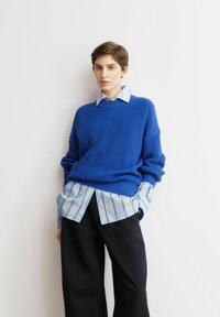 Helles blaues Strickpullover mit lockerem Schnitt, darüber ein hellblau gestreiftes Hemd. Dunkle, weit geschnittene Hosen vervollständigen das Outfit.