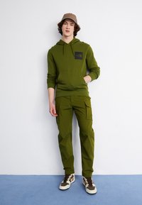 Felpa verde oliva con una toppa a logo nera, abbinata a pantaloni cargo coordinati. Sneakers marroni e bianche completano l'outfit.