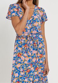 Blumenkleid in Midi-Länge aus blauem Stoff mit rosa, roten und gelben Blumen. Kurze Ärmel, V-Ausschnitt und eine taillierte Detail mit Bindeband und Knöpfen.