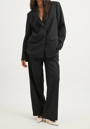 Femme portant un costume noir à fines rayures avec un blazer deux boutons et un pantalon à jambes larges, associé à des talons blancs pointus.