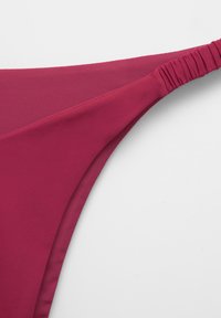 Közeli kép egy magentaszínű bikini alsóról, ráncolt elasztikus oldalszíjjal, világos háttér előtt.